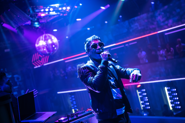 the dj is pictured hear, the lighting around him is magenta, he speaks or signs into a mic and wears aviator sun glasses and a fabulous leather jacket. behind him is a big disco ball. He is having fun. 